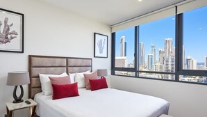 Ein Hotelzimmer mit einem großen Bett, einem Nachttisch, einer Lampe und Blick auf eine Stadtlandschaft durch große Fenster.
