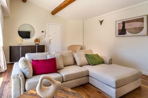 A modern living room with a sectional sofa, a wooden coffee table, a decorative mirror, a wall-mounted artwork, and a door with a light switch.