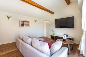 Un salon moderne comprenant un canapé d’angle, une table basse ronde en bois, un téléviseur à écran plat et un coin repas avec une petite table et des chaises.