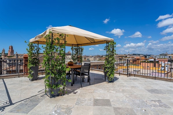 A rooftop terrace with a table and chairs under a canopy, surrounded by potted plants.
