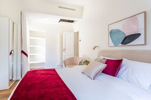 Ein Schlafzimmer mit Bett, Kissen, einer roten Tagesdecke, einem Sessel, einem Nachttisch und einem Bild an der Wand.