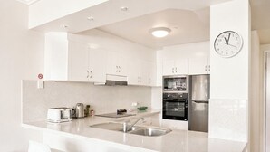 A modern kitchen with white cabinets, stainless steel appliances, a built-in oven, a refrigerator, a sink, and a clock on the wall. - Crown Towers Stunning 1Bedroom Apt 3-min to Beach (Surfers Paradise)