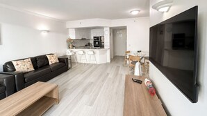A modern living room with a black leather sofa, a wooden coffee table, a TV mounted on the wall, and a kitchen area in the background. - Crown Towers Stunning 1Bedroom Apt 3-min to Beach (Surfers Paradise)