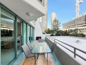 A modern balcony with a glass table and chairs, a view of tall buildings, and a clear sky.