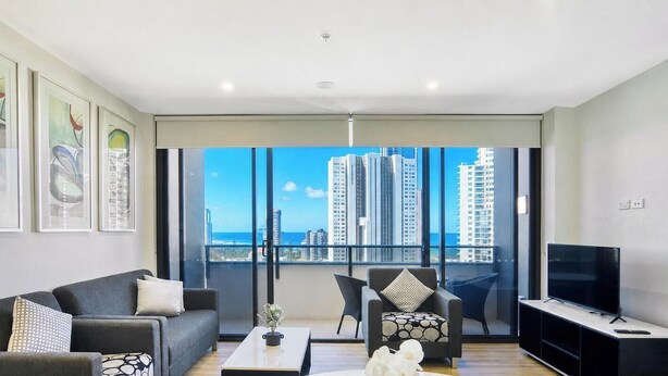 A modern living room with a sofa, armchairs, a coffee table, a TV, and large windows with a city view. - Two Bedroom Apartment with view In SurfersParadise (Surfers Paradise)
