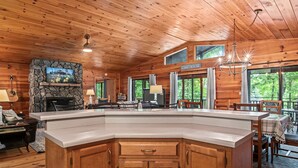 Interior de una cabaña de madera con chimenea de piedra, una isla grande en la cocina y un comedor con vistas al exterior.