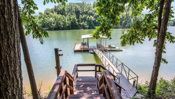 Una terraza de madera que conduce a un muelle con un área de descanso cubierta, rodeada de árboles y un lago tranquilo.