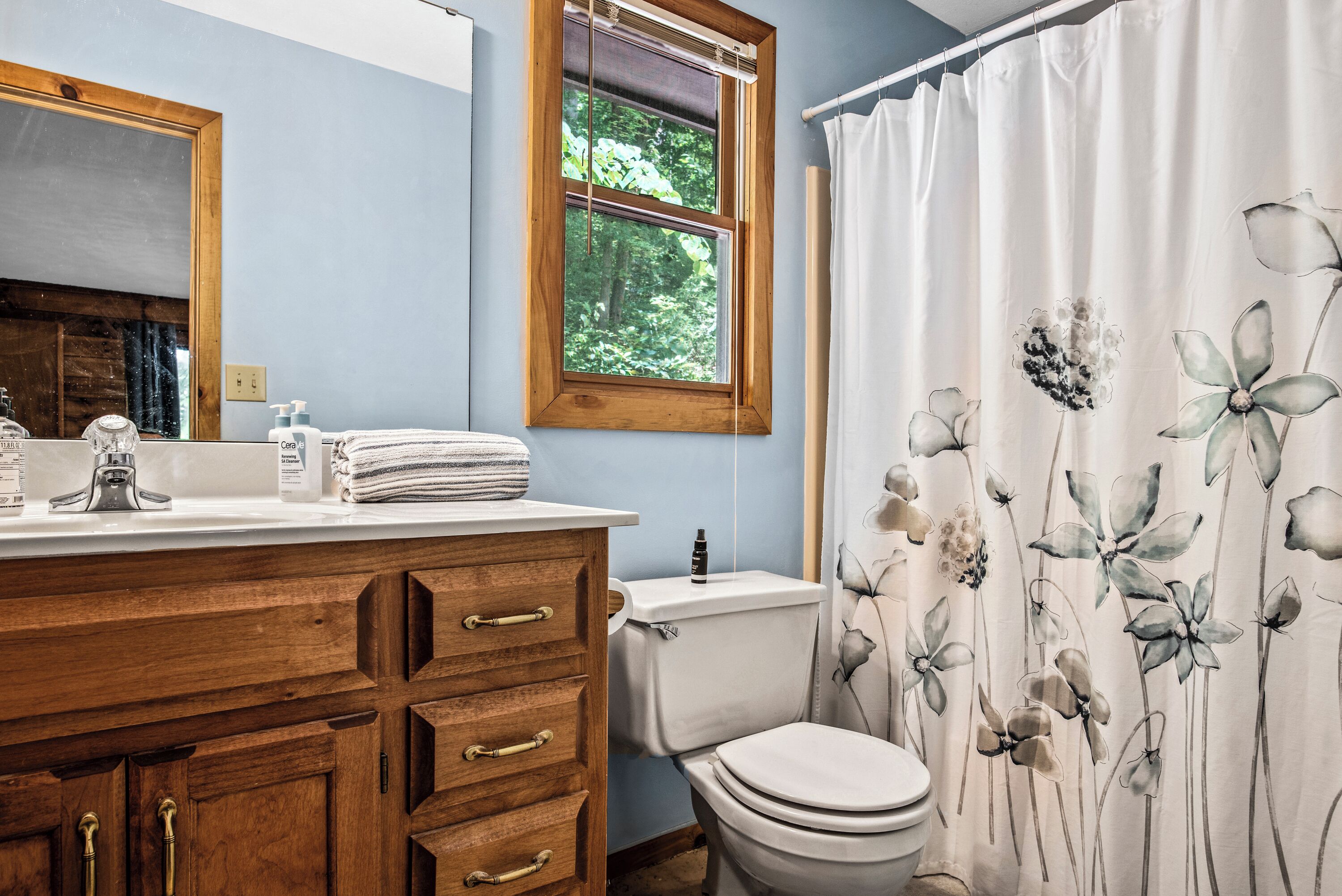 A bathroom with a wooden vanity, a toilet, and a window with a view of greenery.
