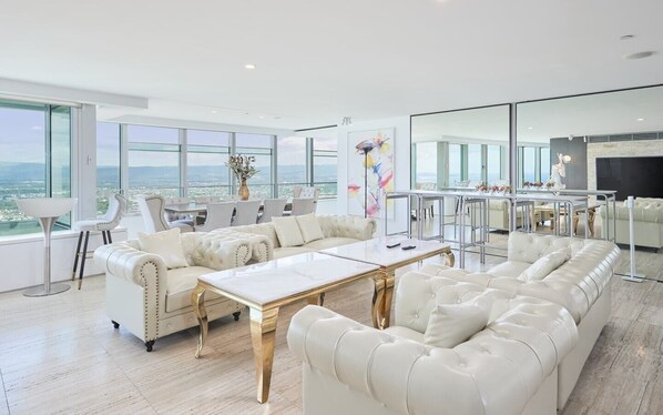 A spacious living room with a large white sofa, a coffee table, and a view of the city through large windows.