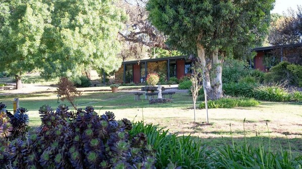 A building with a stone facade, surrounded by a well-maintained garden with various plants and a large tree. - Comfy Studio 3 @ Blickinstal Barossa Valley (Bethany)