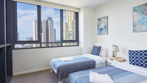 Habitación de hotel con dos camas, un ventanal grande con vista a la ciudad, un cuadro en la pared y una mesita de noche con lámpara.
