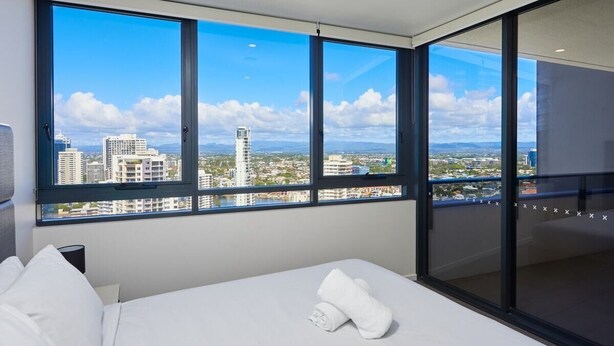 Ein Hotelzimmer mit großem Fenster und Blick auf die Stadt, einem Bett mit weißer Bettwäsche und einer Glastür, die auf einen Balkon führt.