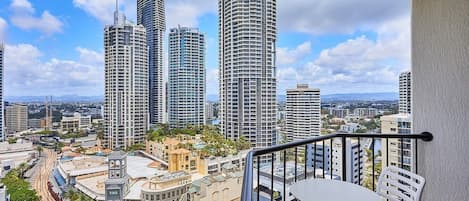 A balcony with a table and chairs, overlooking a cityscape with tall buildings and a clear sky.