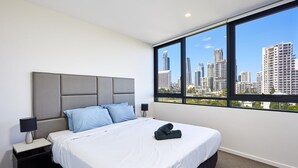 Une chambre d’hôtel moderne avec un grand lit, une tête de lit grise et une vue sur la ville par une grande fenêtre.