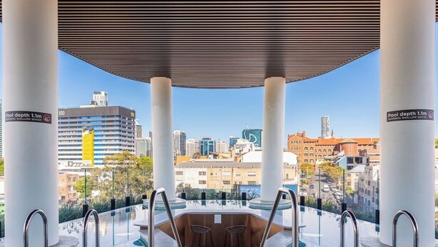 Una piscina infinita al aire libre con vistas a la ciudad, sostenida por columnas blancas, y un letrero de advertencia sobre agua poco profunda.