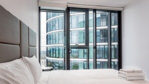 Habitación de hotel moderna con una cama grande, cabecera gris y vistas a un edificio de varios pisos a través de ventanas de piso a techo.