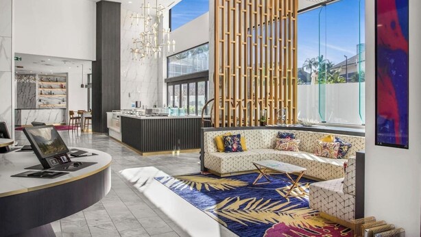 Un moderno vestíbulo de hotel con un mostrador de recepción curvo, una zona de estar con cojines estampados, un panel de madera decorativo y un ventanal con vistas al exterior.