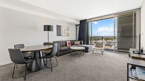 En moderne stue med spisebord, sofa og stole. Der er et stort vindue med persienner, en gulvlampe og et fladskærms-tv på et stativ.