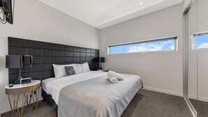 A modern hotel room with a large bed, a bedside table with a lamp, a flat-screen TV mounted on the wall, and a window with a view of the sky.