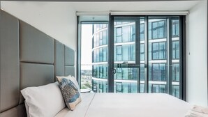 Una habitación de hotel moderna con una cama grande, un cabecero gris y vistas a un paisaje urbano a través de ventanas de gran altura.