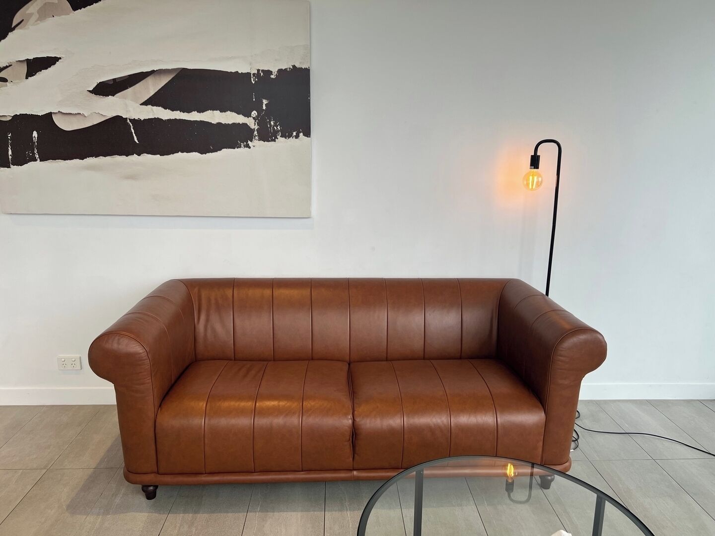 Un canapé en cuir marron dans une pièce dont le mur est blanc et qui est ornée d’un grand tableau abstrait.