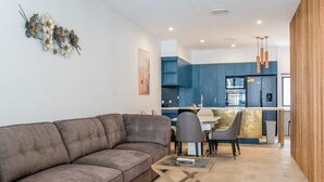 Ein modernes Wohnzimmer mit einer Eckcouch, einem Glastisch und einem Essbereich, im Hintergrund ist eine Küche zu sehen.