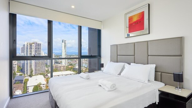 A hotel room with a large bed, a headboard, a nightstand, a lamp, and a window with a city view. - Stylish 1 Bedroom + Partial Ocean View In Surfers (Surfers Paradise)