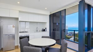 A modern kitchen with a round dining table and grey chairs, a refrigerator, and a view of the ocean through large glass windows. - Stylish 1 Bedroom + Partial Ocean View In Surfers (Surfers Paradise)