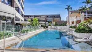 A modern hotel with a rectangular pool surrounded by lounge chairs and palm trees.