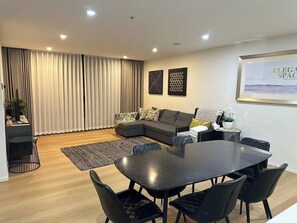 Ein modernes Wohnzimmer mit einer dunklen Couch, einem Esstisch mit Stühlen und einem großen Fenster mit Vorhängen.