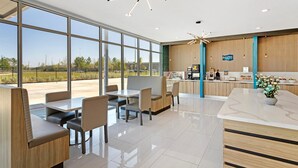 A modern restaurant with a clean, minimalist interior, featuring a central seating area with tables and chairs, a counter with a coffee station, and large windows offering a view of the outdoors. - Clarion Pointe Spring - Houston North (Spring)