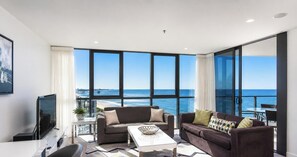 A modern living room with a large window overlooking the ocean, a flat-screen TV, and a dining area with outdoor seating.