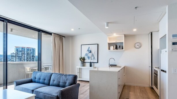 Eine moderne Wohnung mit Wohnbereich, ausgestattet mit einem grauen Sofa und einem Esstisch, einer Küchenzeile mit integriertem Backofen und einem großen Fenster mit Blick auf die Stadt.