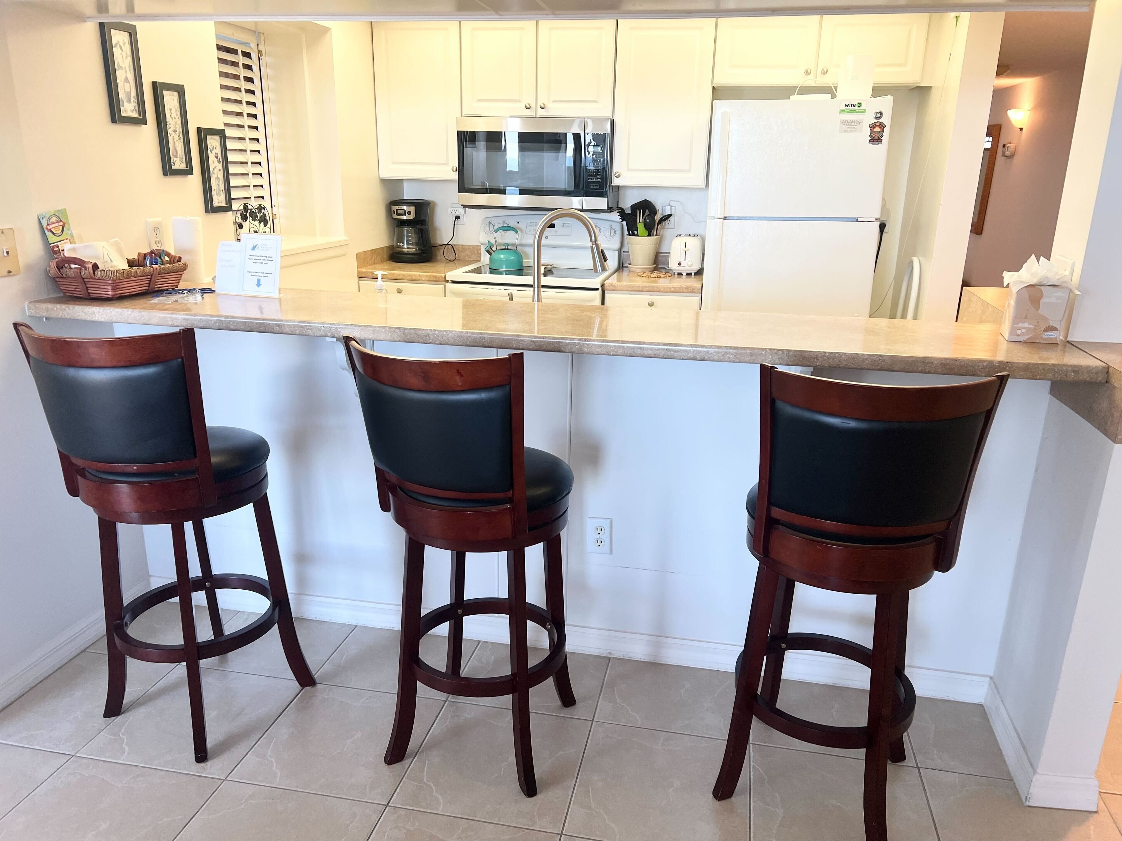 Uma cozinha com ilha central, três banquetas, micro-ondas, geladeira e pia.