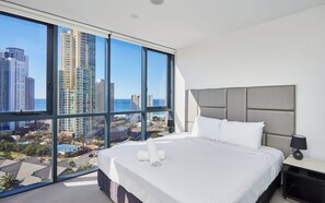 A hotel room with a large bed, a nightstand, and a view of a cityscape through floor-to-ceiling windows.