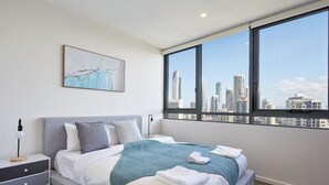 A modern bedroom with a city view, a bed with white linens, a teal throw blanket, and a nightstand with a lamp.