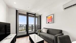 Un salon moderne avec un canapé gris, une table basse et un téléviseur. Une grande fenêtre offre une vue sur la ville.