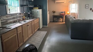 Cozinha compacta com frigorífico, pia e ventoinha. Em área de estar adjacente, vê-se uma mesa de jantar e um sofá.