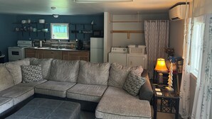 Sala de estar com sofá seccional, uma mesinha com abajur, e área de cozinha com eletrodomésticos e prateleiras.