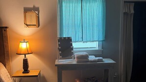 Quarto com cama, uma mesinha com luminária, penteadeira com espelho e iluminação, e janela com cortinas.