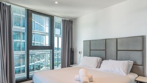Habitación de hotel moderna con una cama grande, cabecero gris y vistas de un paisaje urbano a través de ventanas de piso a techo.