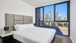 Una habitación de hotel moderna con una cama grande, una mesita de noche, una lámpara y vistas a la ciudad a través de ventanales de piso a techo.