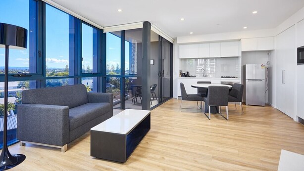 Una sala moderna con un sofá gris, una mesa de centro y un comedor con una mesa y sillas. Hay una zona de cocina con armarios blancos y un refrigerador. Amplias ventanas ofrecen vistas de la ciudad.