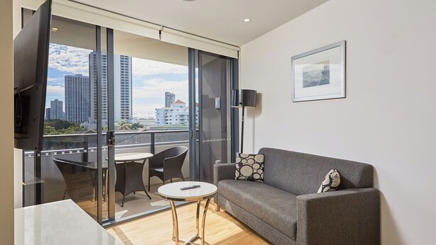 A modern living room with a grey sofa, a small round table, and a glass door leading to a balcony with a city view. - Convenient Located 2-Bedroom Apartment In Surfers (Surfers Paradise)