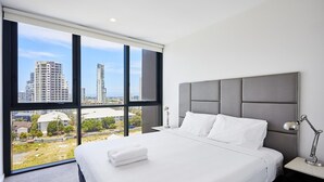 Ein modernes Hotelzimmer mit einem großen Bett, einem grauen Kopfteil und Blick auf hohe Gebäude durch große Fenster.