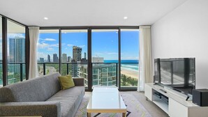 Ein modernes Wohnzimmer mit Sofa, Couchtisch und Fernseher. Es bietet große Fenster mit Blick auf Stadt und Strand.