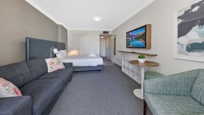 Ein Hotelzimmer mit einem Bett, einem grauen Sofa, einem grünen Sessel, einem kleinen Tisch mit Pflanze und einem an der Wand befestigten Fernseher.