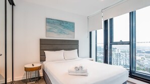 Une chambre d’hôtel moderne avec un grand lit, une table de chevet, un tableau au mur et une vue sur la ville à travers de grandes fenêtres.
