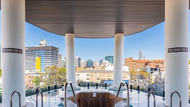 Une piscine extérieure à débordement offrant une vue sur la ville, soutenue par des colonnes blanches, et un panneau d’avertissement indiquant que l’eau est peu profonde.