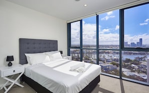 A modern bedroom with a large bed, white bedding, a grey headboard, a nightstand with a lamp, and a view of a cityscape through large windows.